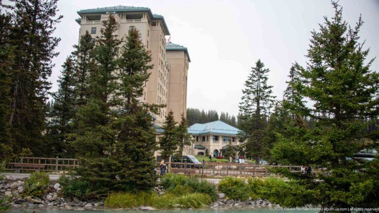 Spruce trees frame the Fairmont Chateau Lake Louise above a road, fence, and creek.