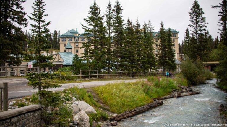 A rushing stream passes a timber bridge below the Fairmont Chateau Lake Louise, partly screened by conifers.