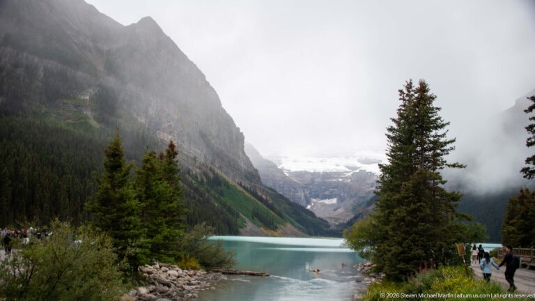 Visitors stand on the rocky shore of Lake Louise as a small boat floats below clouded Victoria Glacier.