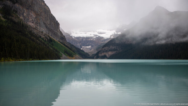 Low cloud partly veils Victoria Glacier above Lake Louise, whose water reflects the mountain valley.