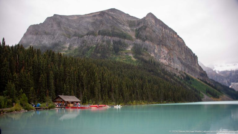 A log boathouse and red canoes sit below a steep mountain slope on Lake Louise.