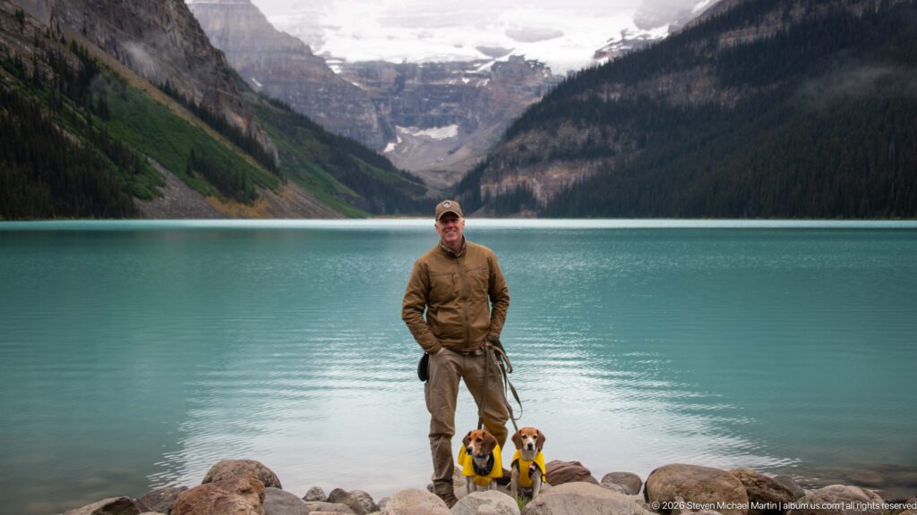 A visitor stands with two leashed beagles on rocks at the edge of Lake Louise.