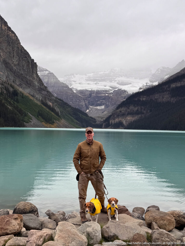 A visitor stands between two yellow-coated beagles on lakeshore stones with the glacier behind them.