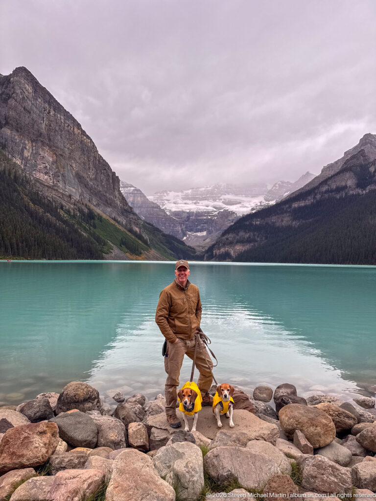 A visitor stands with two yellow-coated beagles on boulders before Lake Louise and the distant glacier.