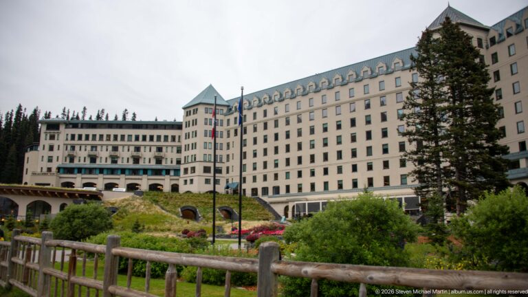 The Fairmont Chateau Lake Louise stands beyond a timber fence and flowered garden, partly screened by conifers.