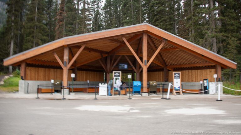 A broad timber shelter with benches and rope queue barriers stands before a conifer forest.