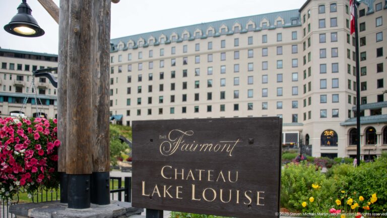 A wooden Fairmont Chateau Lake Louise sign stands among flowers before the hotel façade.