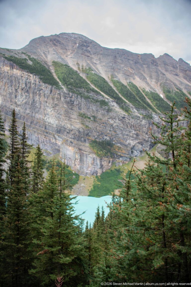 Conifer branches frame a distant turquoise lake below a broad, pale mountain slope.
