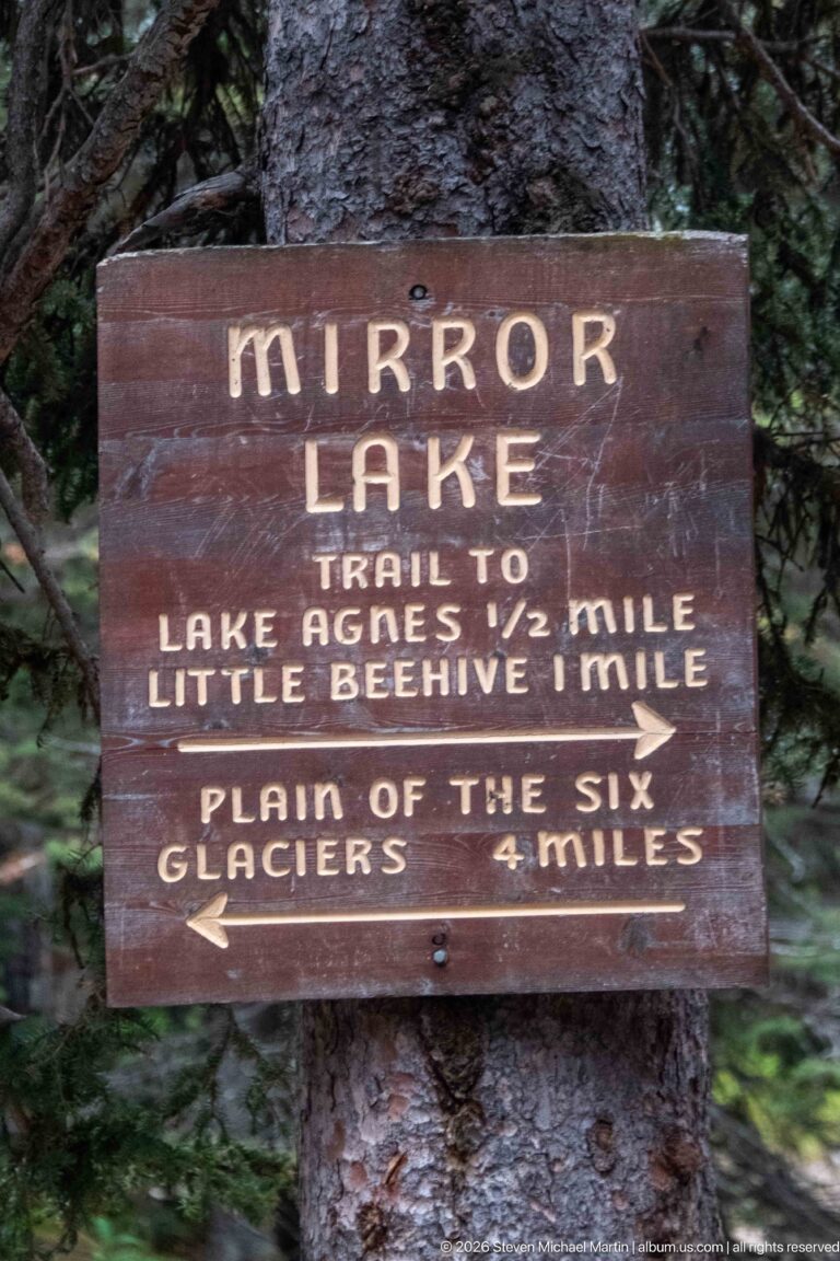 A brown wooden sign gives trail distances from Mirror Lake to Lake Agnes, Little Beehive, and the Plain of the Six Glaciers.