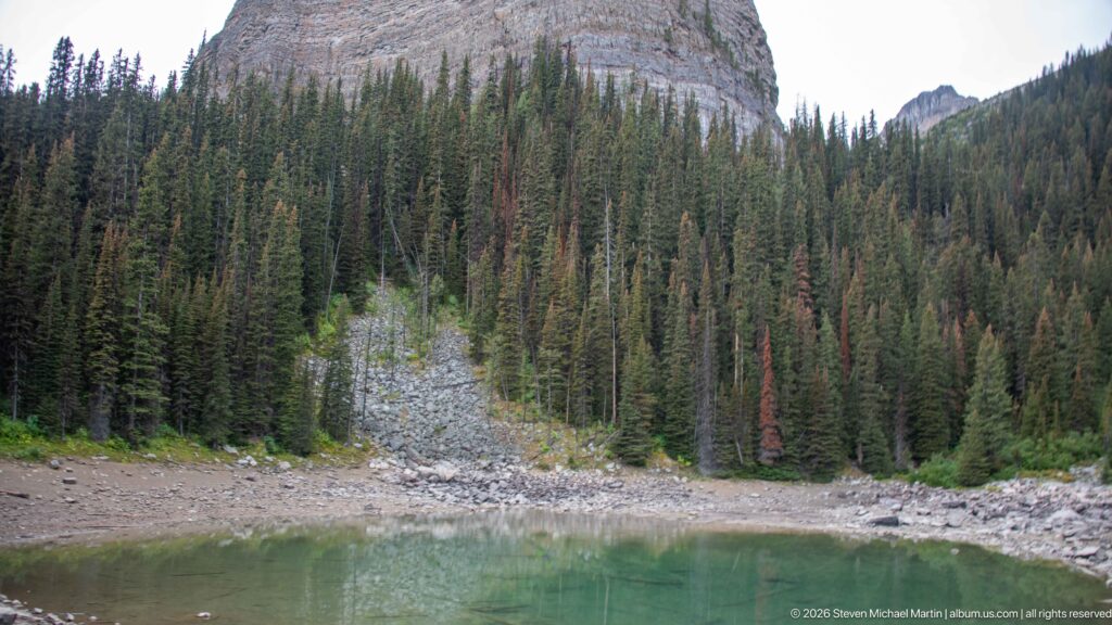 A pale rockslide descends through conifers to the shore of Mirror Lake beneath a rounded cliff.