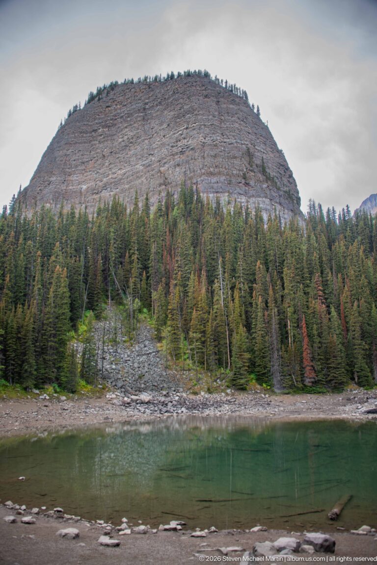 A rounded, layered rock summit rises above Mirror Lake and its stone-strewn shore.