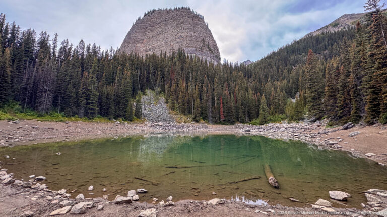 A shallow green lake lies beneath a rounded rock summit, with scree, conifers, and submerged stones.