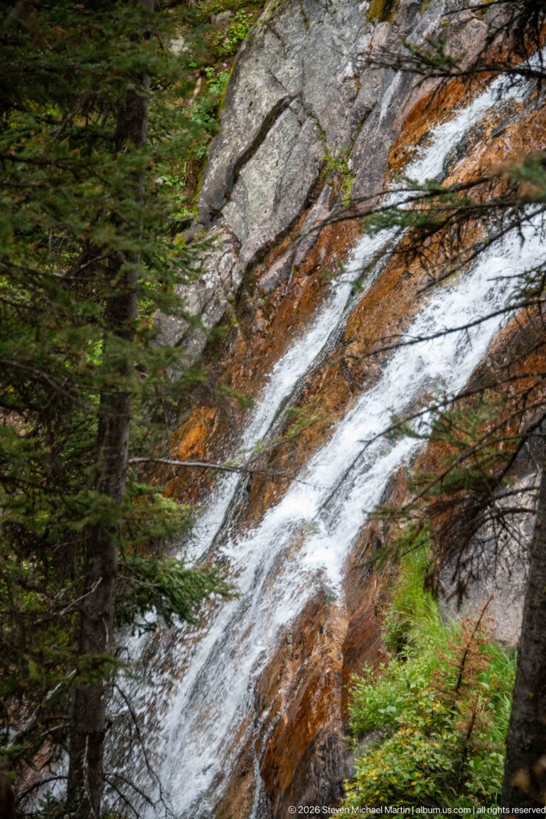 Water slides diagonally over a rust-streaked rock shelf beneath dark evergreen branches.