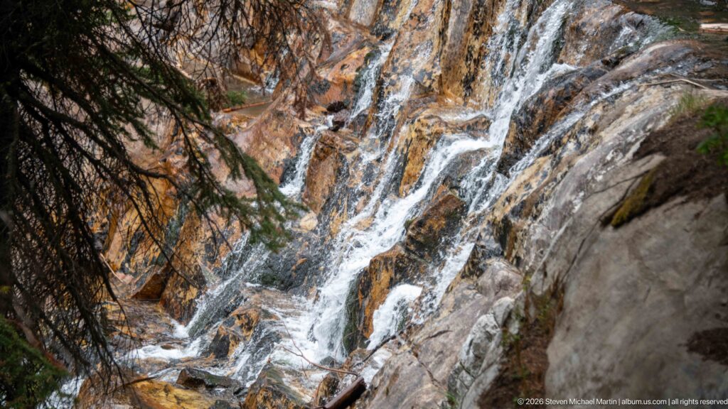 Several narrow streams of white water split across a rust-colored rock face beside dark branches.