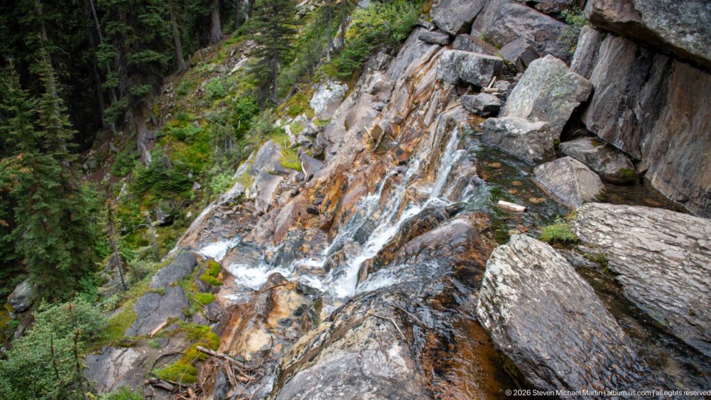 A narrow cascade descends between angular boulders and orange-stained slabs in a forested channel.