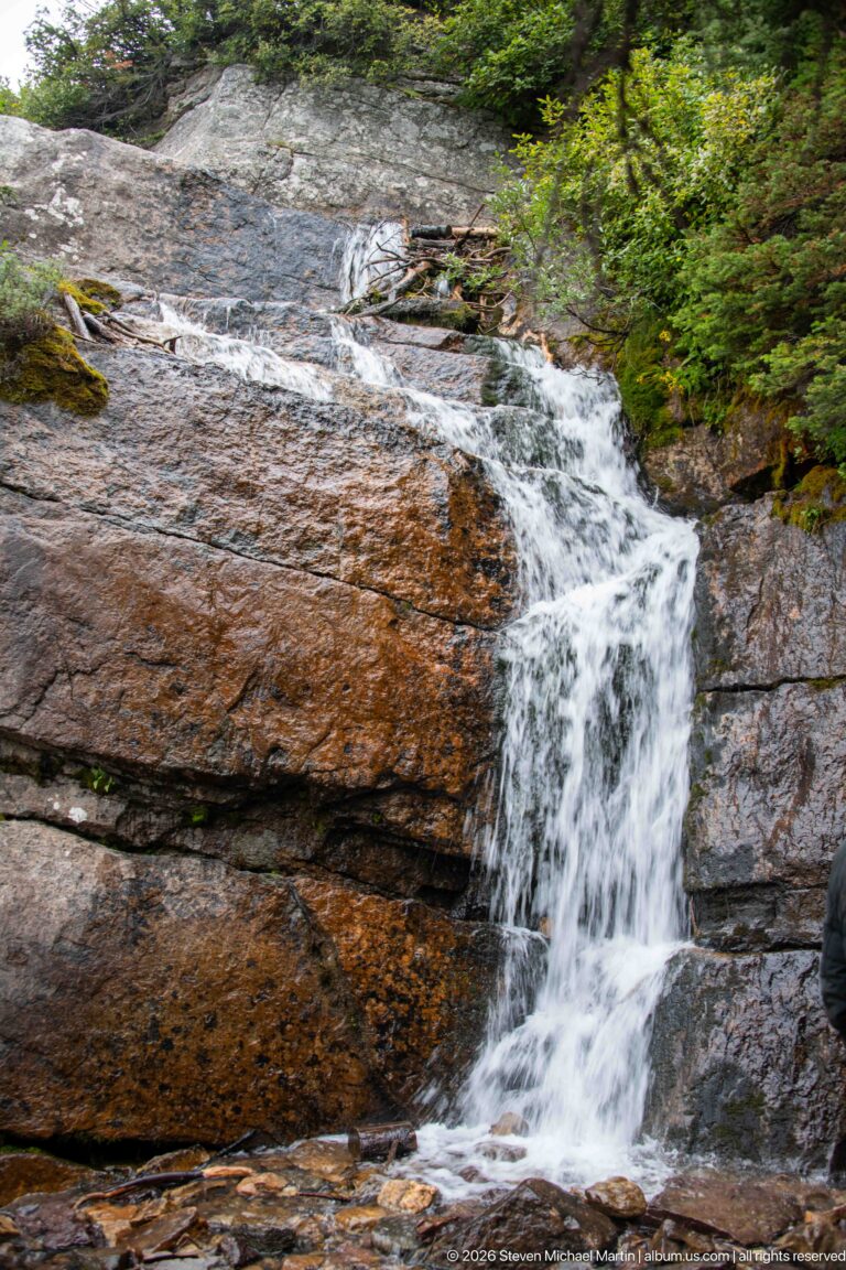 White water falls over rust-colored rock ledges into a dark pool beneath forest branches.