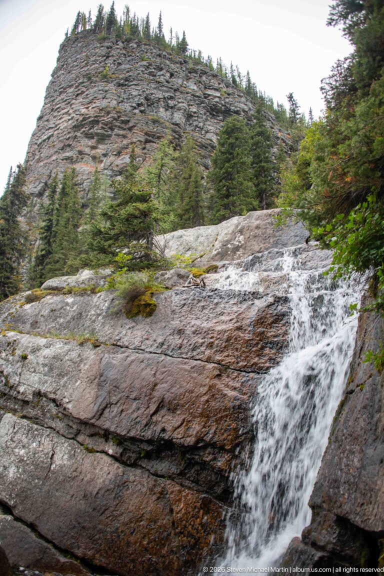 Water runs diagonally over broad gray rock slabs beside conifers and small shrubs.