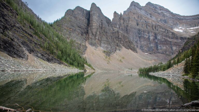 Rock spires and pale scree rise above Lake Agnes and reflect in its still water.