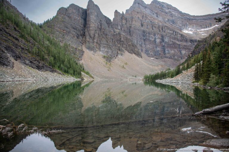 Lake Agnes reflects a steep rocky cirque, with submerged stones visible in the clear near water.