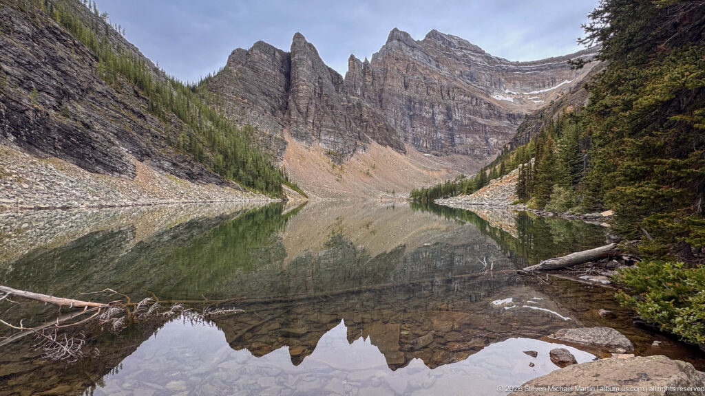 Rock pinnacles and layered cliffs reflect in Lake Agnes, with a conifer slope and log at the shore.