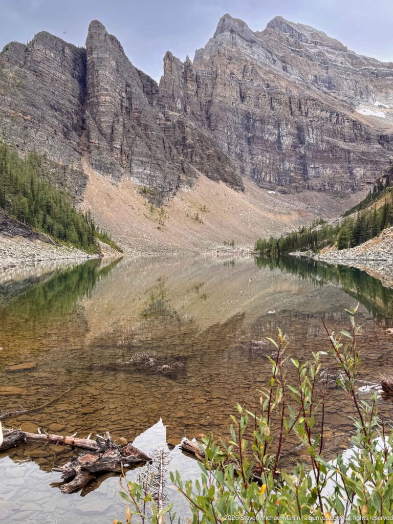 Clear water and driftwood line the near shore of Lake Agnes beneath a broad rocky headwall.