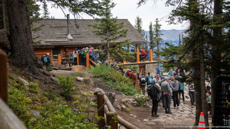 The Lake Agnes Tea House stands among conifers above a stone wall, with hikers gathered at its entrance.