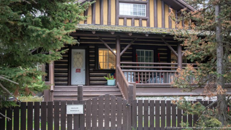 A timber chalet with a covered porch stands behind a private-property fence among spruce trees.