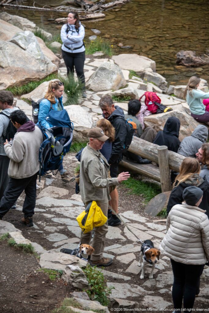 Hikers and two leashed beagles move along the stone path beside a timber rail and lake edge.