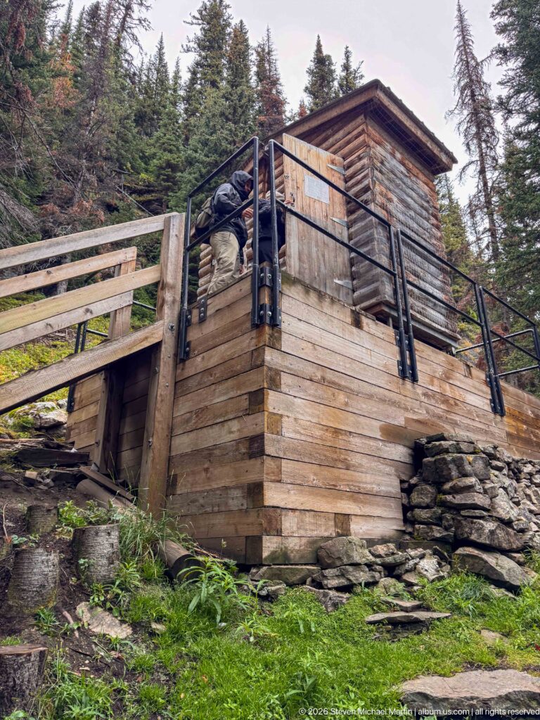 A visitor climbs the exterior stairs of a raised log building on a rocky, forested mountain slope.