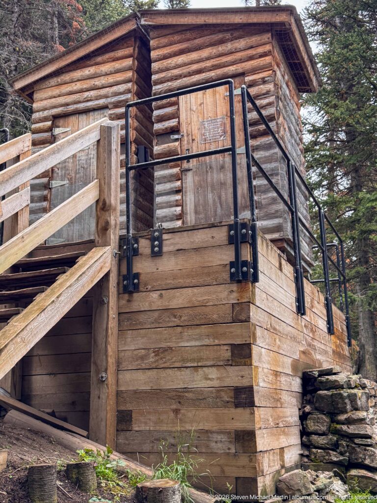 A raised log building with a stone base has exterior stairs and black metal railings beneath conifers.