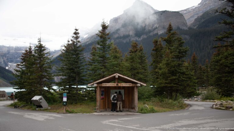 A visitor studies a wooden Lake Louise map kiosk at a paved junction beneath forested mountains.