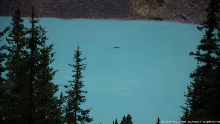 A small red boat crosses a turquoise alpine lake bordered by dark forest and rocky shore.