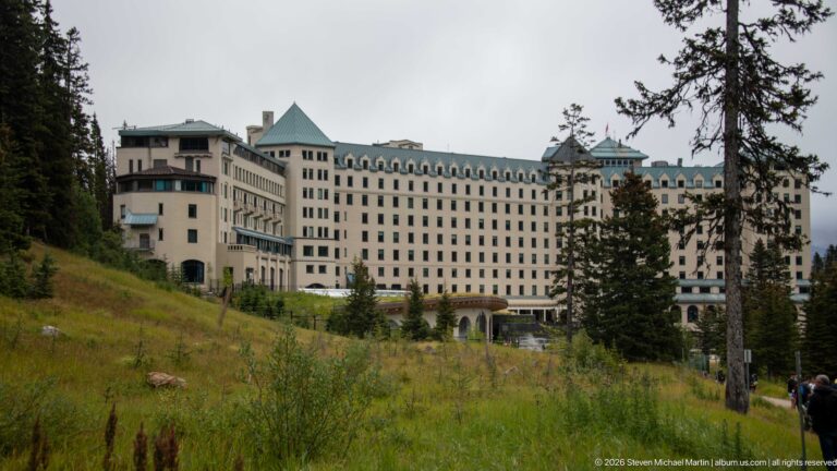 The Fairmont Chateau Lake Louise stands above a rough grassy slope, partly framed by evergreen trees.