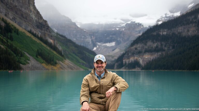 A visitor in a tan jacket sits among shoreline rocks, with Lake Louise and a distant glacier behind him.