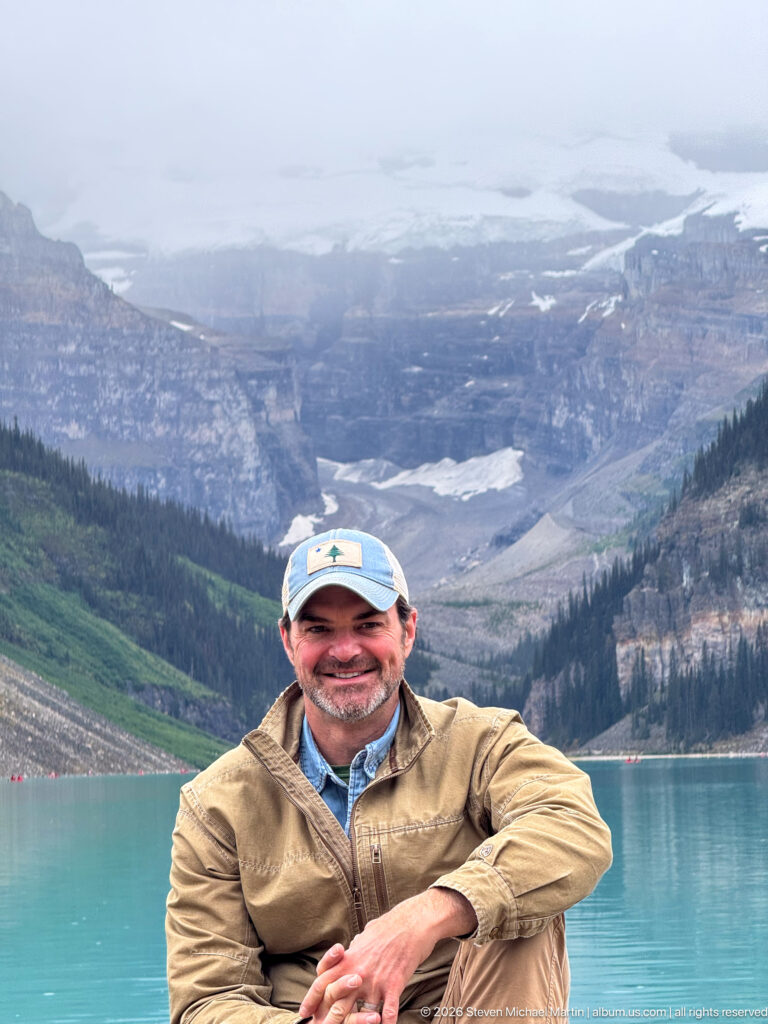 A smiling visitor in a tan jacket and cap sits beside Lake Louise, with the mountain valley softly visible behind him.