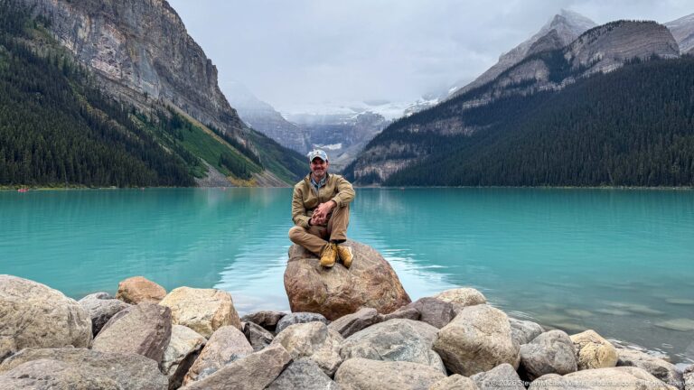 A visitor in a tan jacket sits on a shoreline boulder before turquoise Lake Louise and its mountain valley.