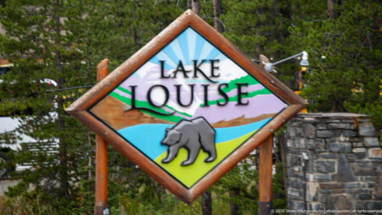 A timber-framed Lake Louise sign shows a painted bear, lake, hills, and sunburst.
