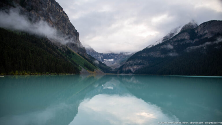 Still turquoise water leads toward a clouded gap between the mountain walls surrounding Lake Louise.