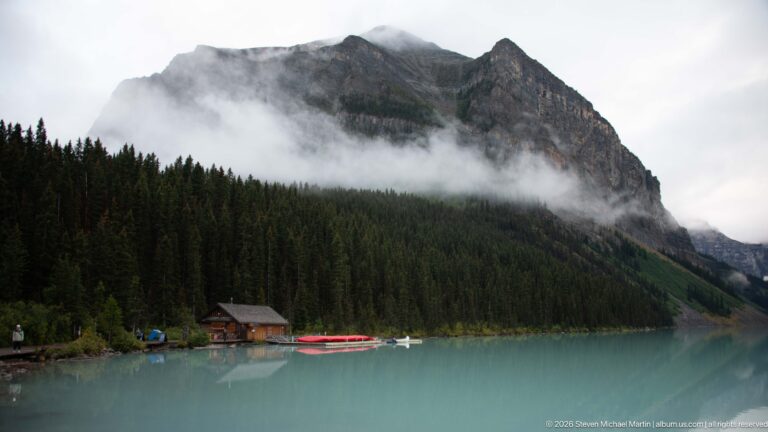 A cloud-wrapped mountain rises above the small log boathouse and red canoes at Lake Louise.