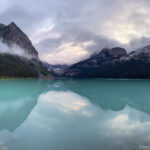 Clouds and steep mountains mirror across Lake Louise, with red canoes visible near the far shore.