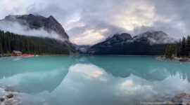 Clouds and steep mountains mirror across Lake Louise, with red canoes visible near the far shore.