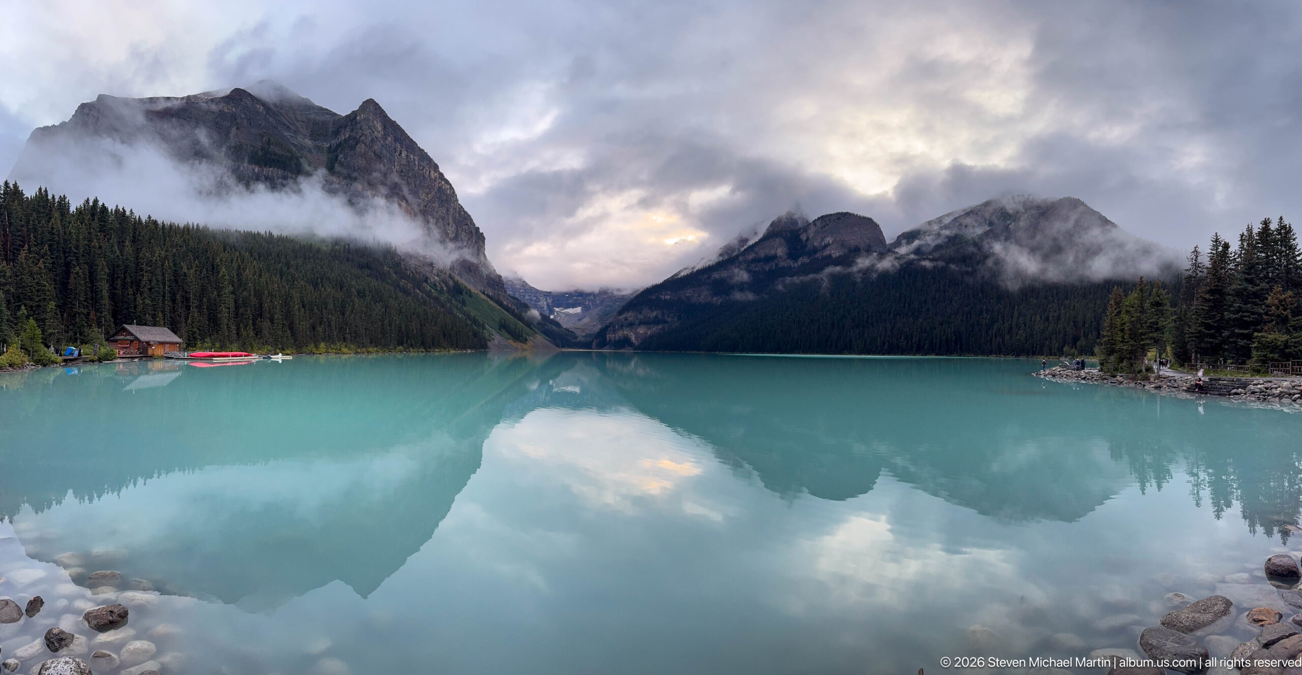 Clouds and steep mountains mirror across Lake Louise, with red canoes visible near the far shore.