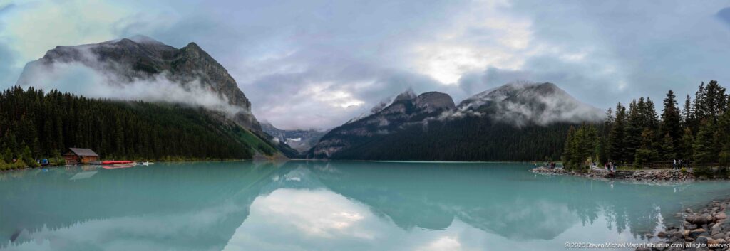 Cloud-wrapped mountain walls and a distant glacier reflect in turquoise Lake Louise beside the far-shore canoe dock.