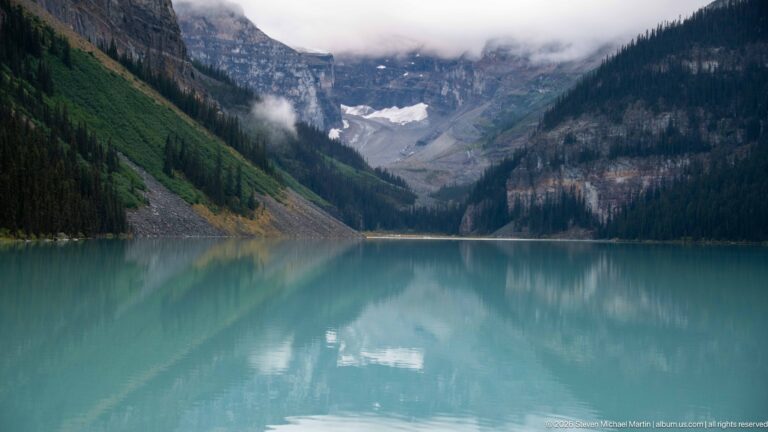A distant glacier lies at the head of Lake Louise between forested mountains, reflected in calm water.