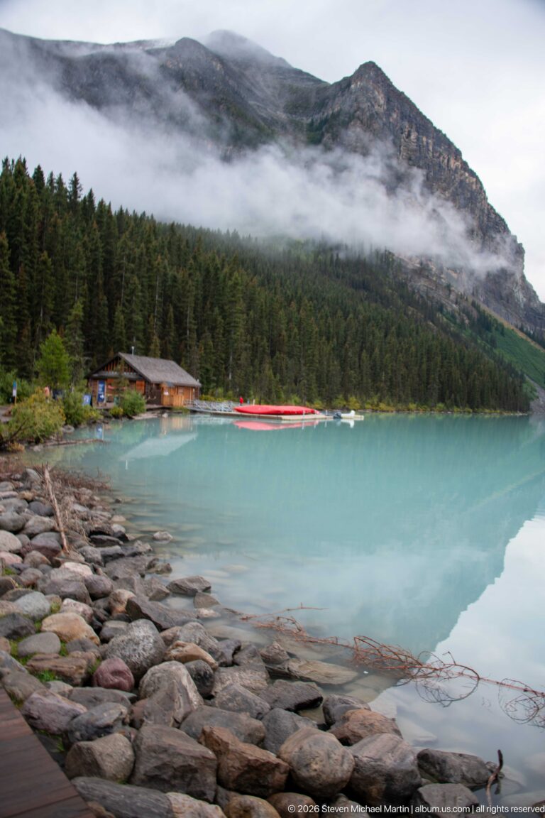 Rounded stones border Lake Louise, with the log boathouse and red canoes across the water.