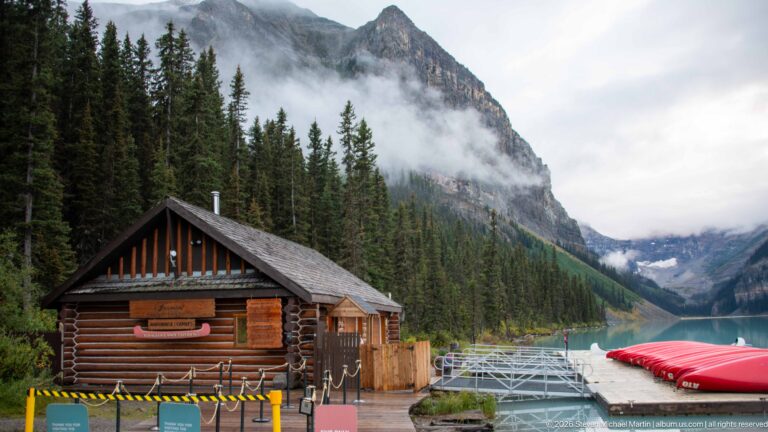 A log boathouse and gangway sit beside red canoes on Lake Louise beneath clouded peaks.