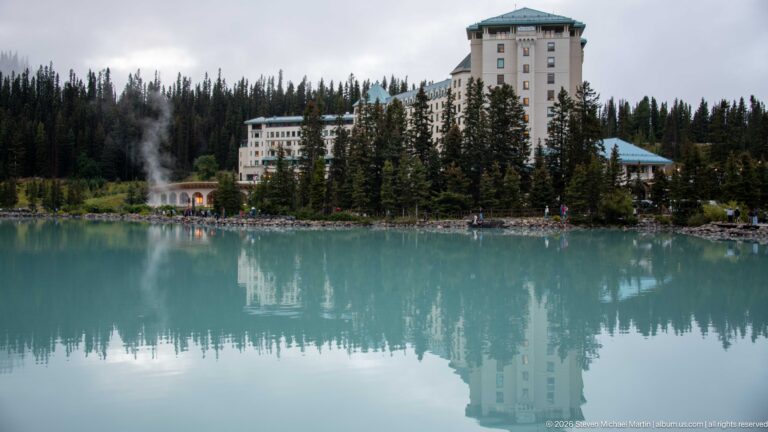 A wide reflection of the Fairmont Chateau Lake Louise stretches across the calm lake below a line of evergreens.