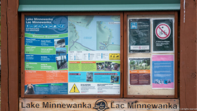 Wooden Lake Minnewanka information kiosk with maps, colored visitor panels, safety notices, and bilingual place name.