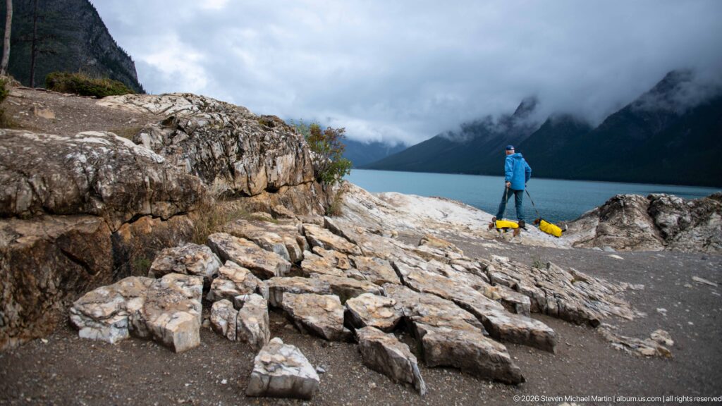 Broken rocky Lake Minnewanka shoreline with a distant visitor and two yellow-coated dogs near the water.