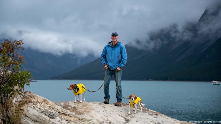 Visitor and two beagles on pale lakeshore rock with Lake Minnewanka and misty mountains behind.